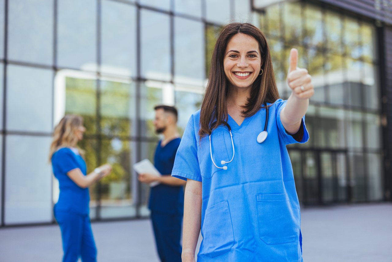 Eine fröhliche Krankenschwester in blauem Kittel zeigt den Daumen nach oben vor einer medizinischen Einrichtung, während ihre Kollegen im Hintergrund ein Gespräch führen.