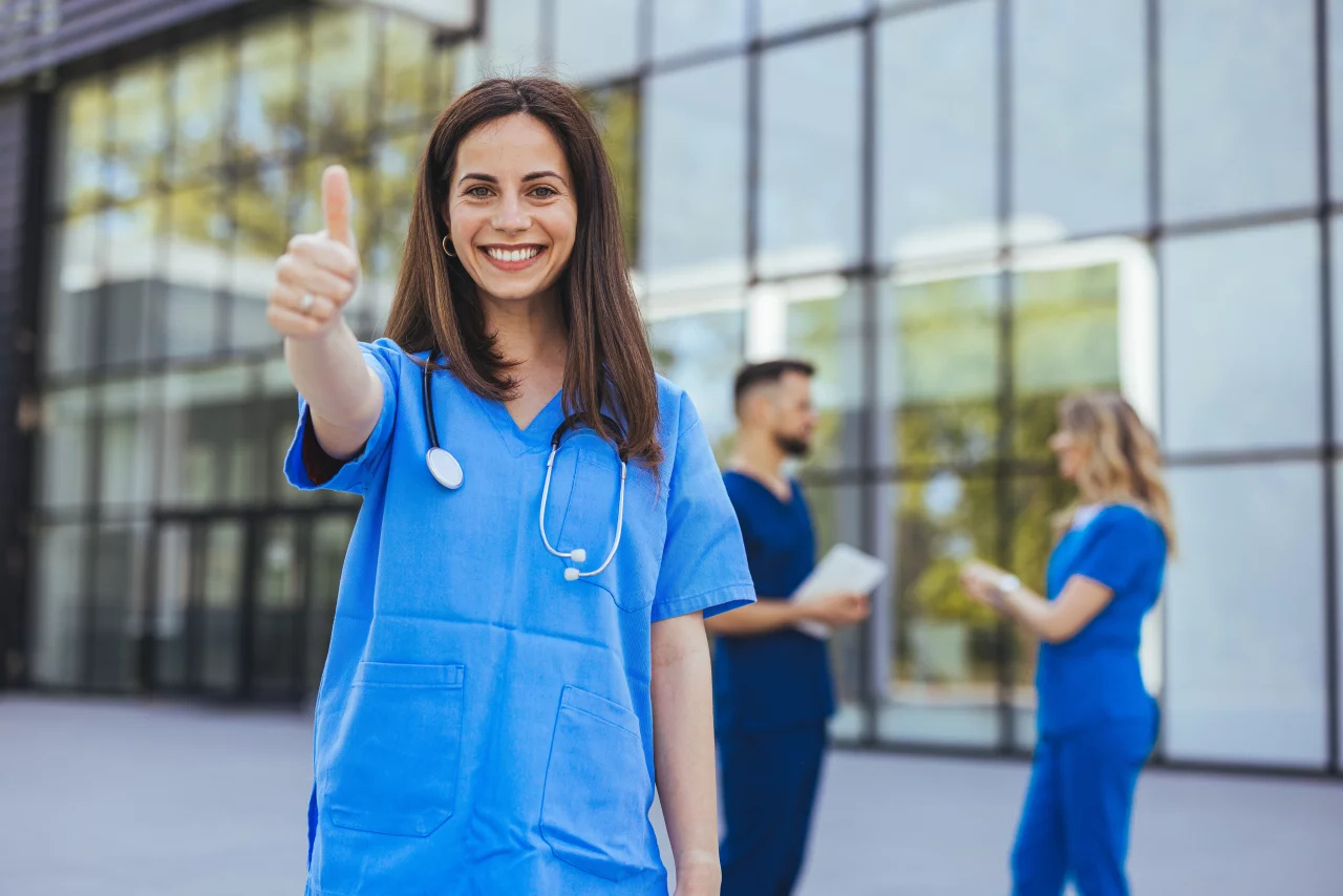 Eine fröhliche Krankenschwester in blauem Kittel zeigt den Daumen nach oben vor einer medizinischen Einrichtung, während ihre Kollegen im Hintergrund ein Gespräch führen.